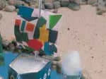 Colorful geometric sailboat near pebbled shoreline on sandy beach.