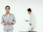 Female professional in suit stands in foreground; blurred colleague walks behind in bright office.