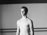 A black and white image of a shirtless male dancer standing in a studio with ballet barres in the background.