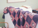 A cozy knitted throw with pink and purple squares draped over a white sofa in a softly lit room.