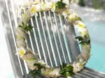 A frangipani and greenery wreath placed on a slatted white wooden chair, with a blurred pool background.