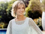 Woman in a crocheted poncho stands outdoors, smiling with blurred greenery and a pool in the background.