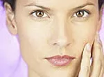 Close-up of a woman's face with clear skin, touching her cheek, against a soft purple background.