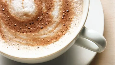 A cup of frothy coffee with cocoa on top, in a white mug on a saucer, placed on a wooden table.