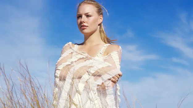A woman stands in tall grass, wrapped in a white shawl, against a clear blue sky.