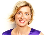 Blonde woman smiling, wearing a purple top, with short hair and dangling earrings, against a white background.