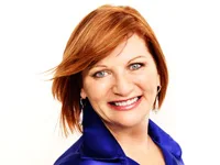 A woman with red hair smiles at the camera, wearing a blue blouse on a white background.