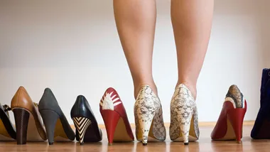 Person standing in snakeskin high heels, surrounded by various other high heeled shoes on a wooden floor.