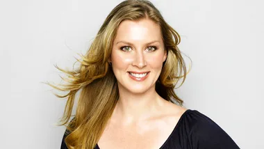 A woman with long blonde hair smiling against a plain background.