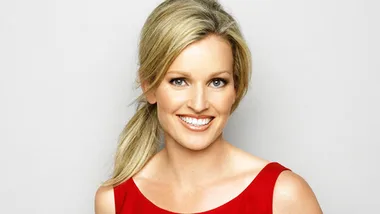 Blonde woman smiling in a red dress, looking towards the camera against a plain background.