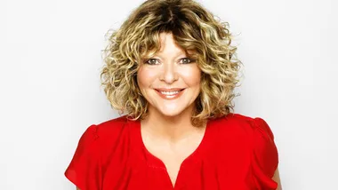A woman with curly hair in a red blouse smiles against a plain background.