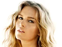 A woman with long, wavy blonde hair looks into the camera against a bright white background.