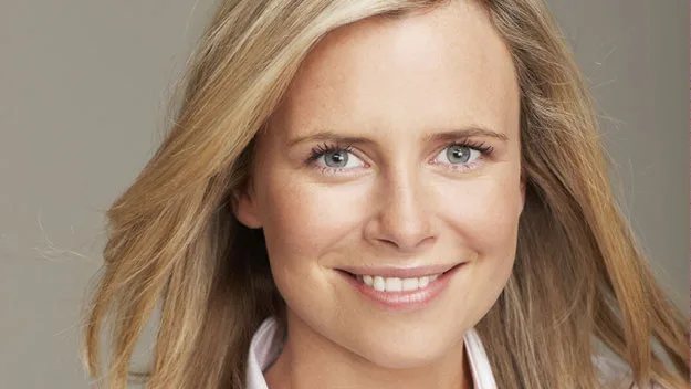 A smiling person with fair skin and long blonde hair, wearing a light-colored top, looking directly at the camera.