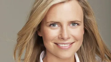 A smiling person with fair skin and long blonde hair, wearing a light-colored top, looking directly at the camera.