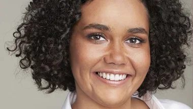 A person with curly hair smiling warmly, wearing a white shirt against a neutral background.