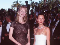 Two women posing on the red carpet at the Cannes Film Festival, with palm trees and people in formal attire in the background.