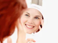 A woman with red hair smiling at herself in a round mirror, set against a bright, white background.