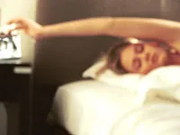 A person lying in bed reaches to turn off an alarm clock on a bedside table.
