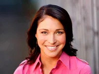 A woman with dark hair smiles warmly, wearing a pink shirt, standing against a blurred background.