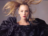 Blonde woman in a dramatic black ruffled outfit, with flowing hair against a grey background.