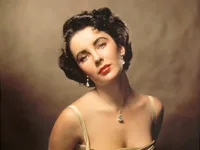 Liz Taylor posing elegantly, wearing a strap dress, with pearl earrings and a necklace, in a classic portrait setting.