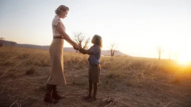 A woman and a child holding hands at sunset on open grassland, with distant hills in the background.