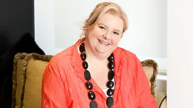 Magda Szubanski smiling, seated on a couch, wearing an orange blouse and large black necklace.