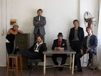 Group of six men in suits and hats pose confidently in a room with a table and a large fan in the background.