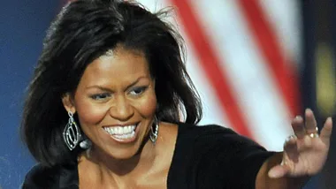 Michelle Obama smiling and waving, wearing earrings, with an American flag in the background.