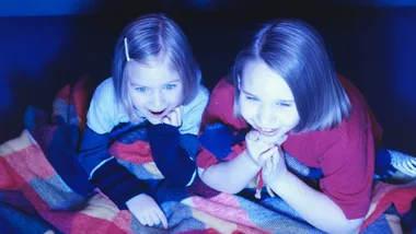 Two children watching TV under a blanket, illuminated by screen light.
