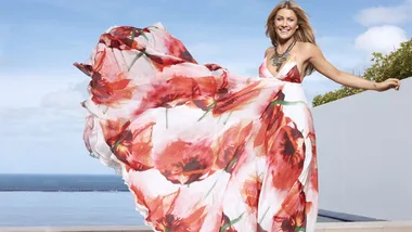 A woman in a flowing floral dress poses joyfully outdoors with the ocean in the background.