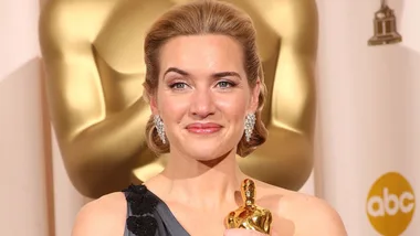A woman smiling while holding an Oscar trophy, standing in front of a large gold statue at an award ceremony.