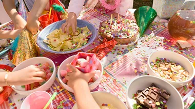Children reaching for snacks and cake on a table decorated with confetti, during a colorful party.