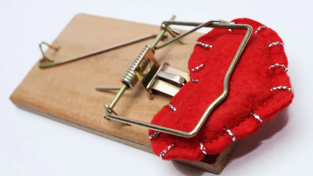 Red felt heart caught in a metal mousetrap on a wooden base, symbolizing emotional pain or heartbreak risk.