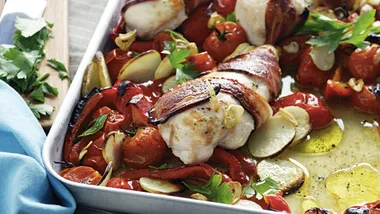 Bacon-wrapped chicken breasts with roasted tomatoes, potatoes, and herbs in a baking dish.