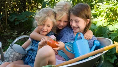 Three children sit in a wheelbarrow, smiling and holding a tomato, surrounded by gardening tools and greenery.