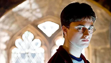 A young wizard with glasses stands in front of an ornate window, bathed in warm light, in a Hogwarts setting.