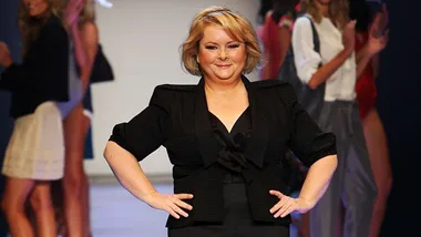 Magda Szubanski confidently walks on a fashion runway, wearing a black outfit, with models in the background.