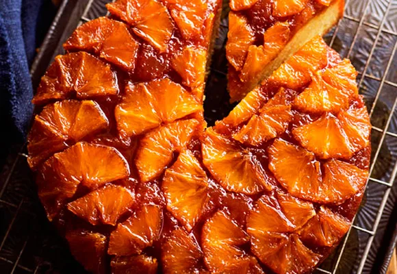 Upside-down cake with caramelized orange slices on top and a slice removed, on a cooling rack.