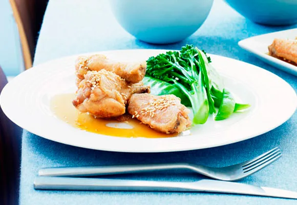 Crispy honey chicken wings with sesame seeds and broccoli on a white plate, placed on a blue tablecloth with cutlery.