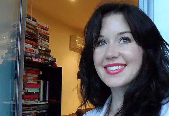 A woman with dark hair smiling inside a room with a tall stack of books behind her.