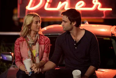 A couple sits on a car holding hands, illuminated by a neon "Diner" sign in the background.
