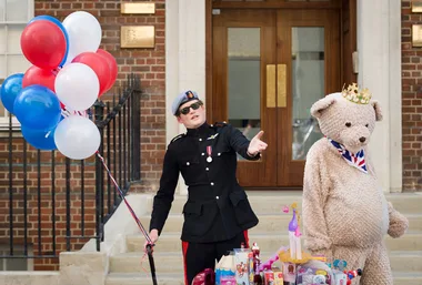 A man in a black uniform and sunglasses holds balloons beside a large teddy bear wearing a crown outside a brick building.