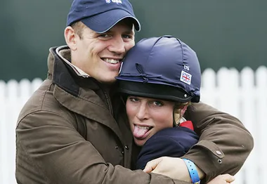 Man in a cap hugging a helmeted woman playfully sticking out her tongue, with a white fence in the background.