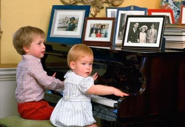 Two young children playing piano, surrounded by framed family photos.