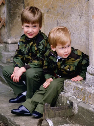 Two young boys in matching camo jackets and green pants sit on stone steps, smiling in an outdoor setting.