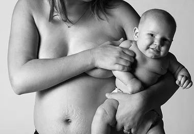 Mother holding baby, both smiling; shows post-pregnancy body with natural stretch marks, promoting body positivity.