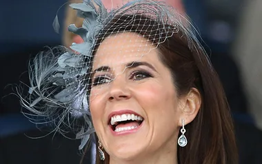 A smiling woman with dark hair wearing a decorative fascinator and teardrop earrings.