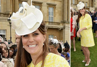 Woman in a yellow coat and white hat smiling at an outdoor gathering with a crowd and historic building in the background.