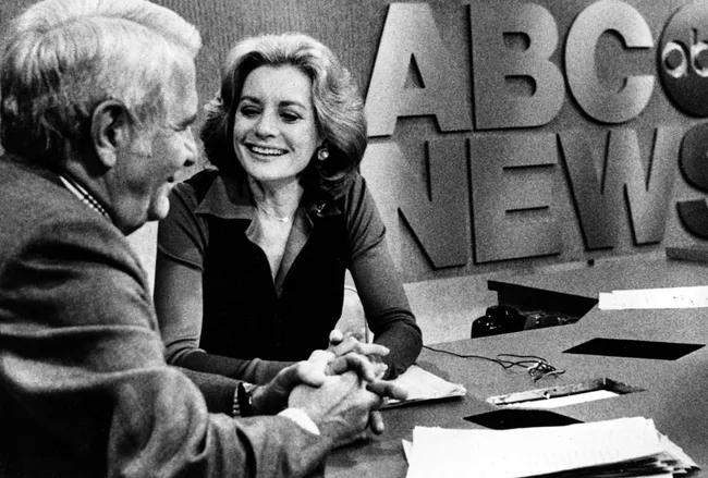 Two people sitting at an ABC News desk, smiling and talking, with papers and a pen on the table.